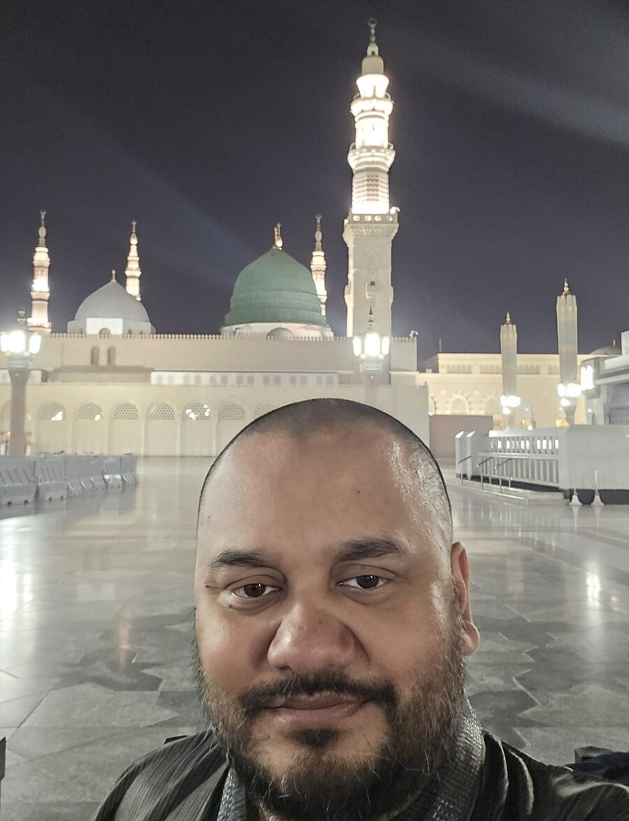Shofiqul Islam standing in the courtyard of Masjid an-Nabawi in Medina at night, with the green dome and minarets illuminated in the background